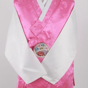 Może przedstawiać: Tradycyjny koreański hanbok w kolorze różowym i białym z haftem kwiatowym. Hanbok ma biały kołnierzyk i długie rękawy. Rękawy są białe z subtelnym wzorem kwiatowym. Hanbok jest noszony na brązowych spodniach.