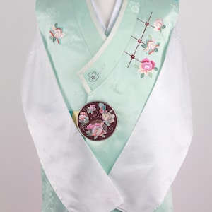 Może przedstawiać: Tradycyjny koreański strój hanbok. Miętowa kamizelka zdobiona jest haftem kwiatowym i okrągłą broszką z kwiatowymi wzorami. Białe rękawy i różowa spódnica dopełniają strój, prezentowany na manekinie.