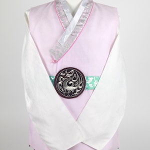 Może przedstawiać: Tradycyjny koreański hanbok w kolorze różowym i białym z kołnierzem w srebrne i różowe wzory. Hanbok ma białą szarfę z zielonym i srebrnym paskiem oraz okrągłą, ozdobną klamrę w kolorze srebrnym i czarnym.