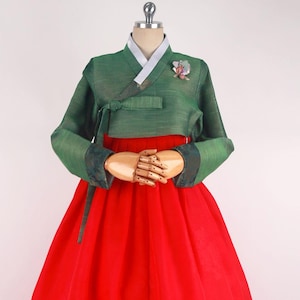 Może przedstawiać: Tradycyjna koreańska suknia hanbok z zielonym gorsetem i czerwoną spódnicą. Gorset ma długie rękawy i ozdobny węzeł w talii. Spódnica jest pełna i falująca.