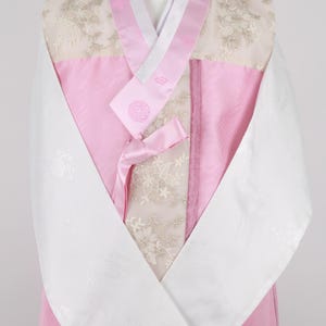 Może przedstawiać: Tradycyjny koreański hanbok w kolorze różowym i białym z białym kołnierzykiem i długimi rękawami. Hanbok ma kwiatowy wzór i różową wstążkę.