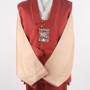 Może przedstawiać: Tradycyjny koreański hanbok w kolorze czerwonym i beżowym z srebrną ozdobą. Hanbok ma długą, spływającą spódnicę i dopasowany gorset z wysokim kołnierzem.