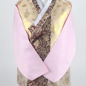 Może przedstawiać: Tradycyjny koreański hanbok, długa,  ubranie z długimi rękawami z różowego materiału z  złotym wzorem. Hanbok ma burgundową wstążkę i biały kołnierzyk.