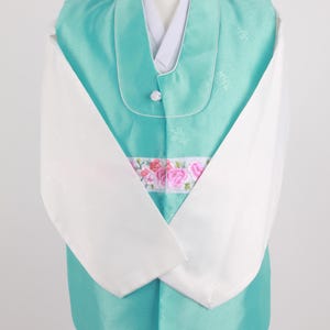 Może przedstawiać: Tradycyjny koreański strój, czyli hanbok, w jasnoturkusowym kolorze z białymi akcentami. Hanbok składa się z długiej kurtki z kwiatowym haftem i białym kołnierzem.