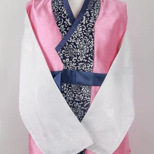 Może przedstawiać: Tradycyjny koreański hanbok w kolorze różowym i białym z niebieskim i białym wzorem kwiatowym. Hanbok ma długie rękawy i pasek.