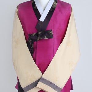 Może przedstawiać: Tradycyjny koreański strój Hanbok. Zestaw zawiera żywą, magentową kamizelkę z białym kołnierzem i czarnym wykończeniem. Rękawy są kremowe ze wzorami kwiatowymi i szarym wykończeniem. Strój jest prezentowany na manekinie.