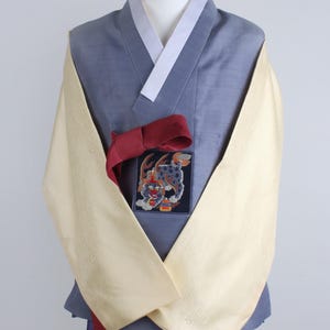 Może przedstawiać: Tradycyjny koreański hanbok prezentowany na manekinie. Zestaw zawiera jasno niebieską kurtkę z białym kołnierzem, kremowymi rękawami i ciemnoczerwoną szarfą. Na piersi znajduje się haftowana naszywka z kolorowym wzorem. Dolna część garderoby to ciemnoniebieska spódnica.