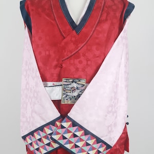 Może przedstawiać: Tradycyjna koreańska szata w kolorze czerwonym i różowym z geometrycznym wzorem na rękawach i białym haftem na przodzie. Szata ma czarny kołnierz i czarny pas.