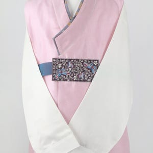 Może przedstawiać: Tradycyjny koreański hanbok w kolorze różowym i białym z haftowaną kwiatową wstążką. Hanbok ma długie rękawy i dekolt w kształcie litery V.