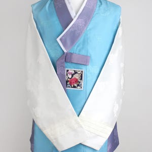 Może przedstawiać: Tradycyjny koreański strój Hanbok, z jasnoniebieską kamizelką i fioletowymi wykończeniami. Rękawy są białe z kwiatowym wzorem. Mała kwadratowa naszywka z kwiatowym wzorem znajduje się z przodu.