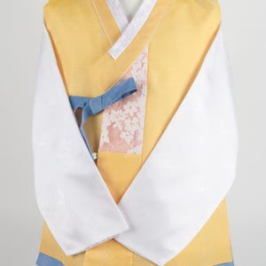 Może przedstawiać: Żółty hanbok z białym kołnierzykiem i długimi rękawami. Hanbok ma niebieską wstążkę zawiązaną w talii i różowo-biały wzór kwiatowy z przodu. Hanbok jest noszony na ciemnoniebieskich spodniach.