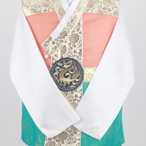 Może przedstawiać: Tradycyjny koreański hanbok, długa koszula z kolorowym wzorem. Koszula jest w kolorze zielonym, brzoskwiniowym i białym z złotymi akcentami. Na przodzie odzieży widoczny jest okrągły emblemat ze złotym wzorem feniksa.