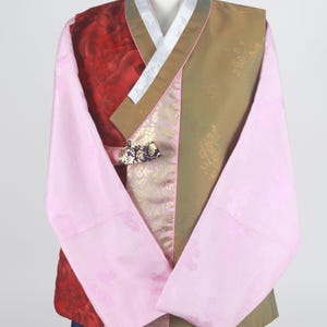 Może przedstawiać: Tradycyjny koreański hanbok, długa koszula z rękawami, w różowym, czerwonym i złotym wzorze. Hanbok ma biały kołnierz i złotą wstążkę.