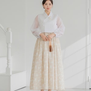 Op de afbeelding: Een vrouw draagt een traditionele Koreaanse Hanbok. De outfit bestaat uit een witte top met doorschijnende mouwen en een beige rok met een bloemenpatroon. De outfit wordt gecompleteerd met beige hakken en een klein accessoire.