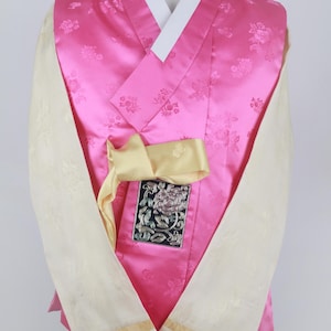 Może przedstawiać: Różowy hanbok z kwiatowym wzorem, białym kołnierzykiem i żółtą wstążką. Hanbok ma ozdobną haftowaną łatę z przodu.