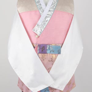 Może przedstawiać: Tradycyjny koreański hanbok, długa bluzka z różowo-beżowym gorsetem, białymi rękawami i kolorową wzorzystą wstążką. Hanbok jest noszony na brązowej spódnicy.