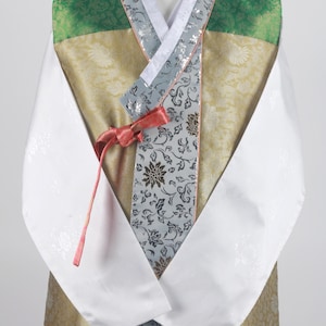 Może przedstawiać: Tradycyjny koreański hanbok, długa szata z długimi rękawami, białym kołnierzem i wzorem w kolorze zielonym, złotym i srebrnym. Hanbok jest związany czerwoną wstążką.