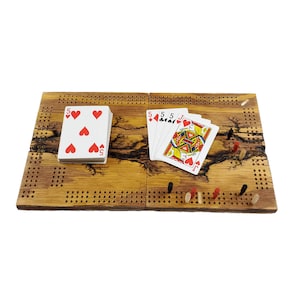 May include: A wooden cribbage board with a natural wood finish and black and red pegs. The board is divided into two sections, each with a cribbage board layout. There are playing cards on the board, including a five of hearts and a jack of hearts.
