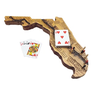 May include: A wooden cribbage board shaped like the state of Florida. The board has a burnt wood finish and is made of multiple layers of wood. The board is shown with playing cards and pegs.