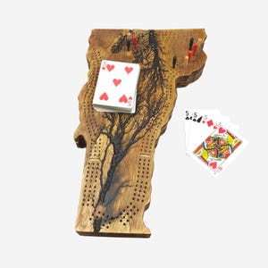 May include: A wooden cribbage board shaped like the state of Vermont. The board has a burnt wood design and is decorated with a tree. There are playing cards and pegs on the board.