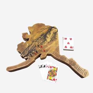 May include: A wooden game board shaped like the state of Alaska with a black and white tree design. The board has a grid of holes and red and black pegs are placed in some of the holes. There are also playing cards on the board, including a Jack of Hearts and five of a kind of fives.