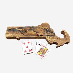 May include: A wooden cribbage board shaped like the state of Massachusetts. The board has a dark brown wood grain and a black burnt wood design. There are five cards on the board, including a five of hearts, a five of spades, a five of clubs, a jack of diamonds, and a queen of hearts.