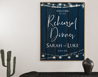 Pode incluir: Uma placa de madeira azul com texto branco que diz "Bem-vindos ao nosso jantar de ensaio Sarah e Luke 14 de julho de 2022". A placa é decorada com luzes brancas.