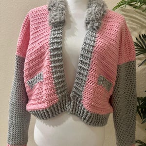 May include: A handmade, open-front crochet jacket in pink and gray, with a faux fur collar. The sleeves and trim are gray, while the body is pink. The jacket is displayed on a white mannequin.