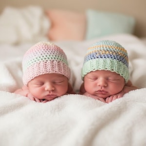 May include: Two newborn babies wearing pastel-colored crocheted hats, resting on a soft, white blanket. One hat is pink, the other is green, with stripes of blue, yellow, and orange. The babies are peacefully asleep.