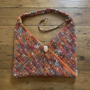 May include: A handmade, colorful crochet bag with a braided strap and a detachable strap. The bag features a mix of orange, pink, teal, and brown yarns. It has a leather closure and gold-colored hardware.