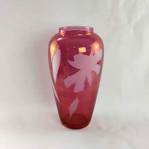 May include: A tall, ruby-red glass vase with a floral design. The vase features a large etched flower and leaf pattern in a lighter pink hue. The vase has a rounded body and a flared rim, suitable for displaying flowers.