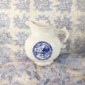 May include: White ceramic pitcher with a blue and white Asian-inspired design. The pitcher has a rounded body, a spout, and a handle with a decorative swirl. The design is a circular medallion with a landscape scene.