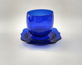 May include: A cobalt blue glass cup and saucer set. The cup is a rounded shape, and the saucer has a scalloped edge. The set is made of a transparent, glossy glass material. The set is on a white background.