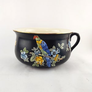 May include: A black ceramic bowl with a handle, featuring a colorful parrot perched on a branch with blue and yellow flowers. The rim is cream-colored with a gold trim. The bowl has a wide opening and a curved shape.