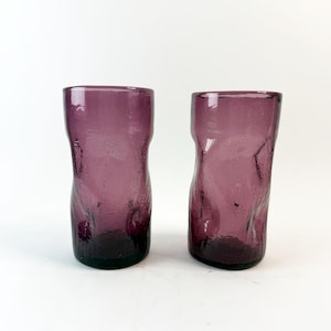Puede incluir: Dos vasos morados con un diseño texturizado y orgánico. Los vasos tienen una base oscura y un cuerpo más claro y translúcido. La forma es ligeramente irregular, lo que añade a su estética única.