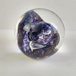 May include: A clear glass paperweight with a swirling, abstract design in shades of purple and black. The interior features a textured, organic shape with a speckled pattern, encased within the smooth, spherical glass.