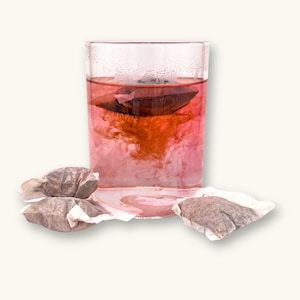 May include: A clear glass of pink tea with a tea bag steeping inside. The tea is a deep pink color, and the tea bag is dark brown. Several unused tea bags are scattered around the glass.