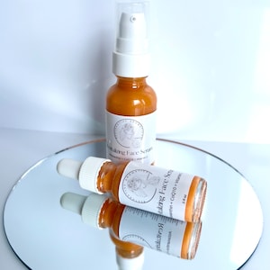 May include: Two amber glass bottles with gold lids and droppers. The bottles are labeled "Rejuvenating Face Serum" with a list of ingredients including CoQ10 and Vitamin C. The bottles are sitting on a silver surface.