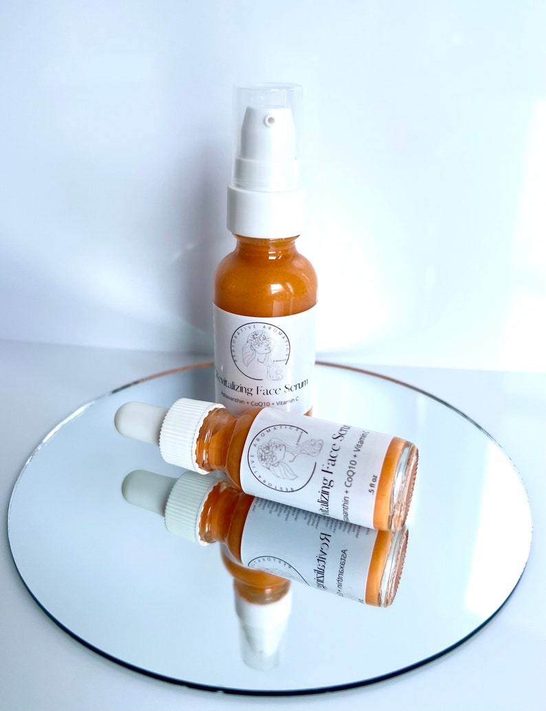 May include: Two amber glass bottles with gold lids and droppers. The bottles are labeled "Rejuvenating Face Serum" with a list of ingredients including CoQ10 and Vitamin C. The bottles are sitting on a silver surface.