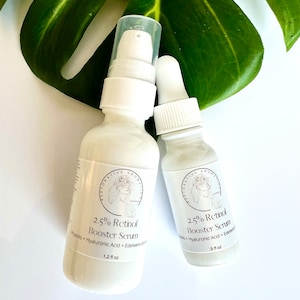 May include: Two white bottles of 2.5% Retinol Booster Serum with a white dropper top. The bottles are labeled with the product name and ingredients, including Propolis, Hyaluronic Acid, and Edelweiss Extract. The bottles are sitting on a white surface with a green leaf in the background.