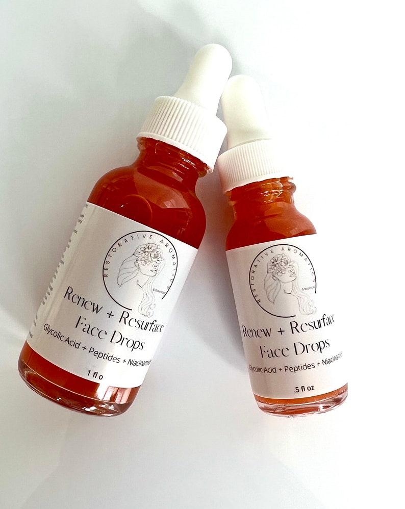 Może przedstawiać: Dwie bursztynowe butelki ze szklanymi kroplomierzami i białymi etykietami. Etykiety zawierają czarno-biały rysunek twarzy kobiety z kwiatami we włosach. Tekst na etykietach brzmi "Restorative Aromatics" i "Renew + Resurface Face Drops". Mniejsza butelka jest oznaczona "1 fl oz", a większa "5 fl oz".