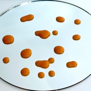 May include: A close-up of a round, silver mirror with multiple small, orange-brown drops of liquid scattered across the surface.