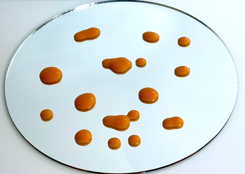 May include: A close-up of a round, silver mirror with multiple small, orange-brown drops of liquid scattered across the surface.