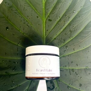May include: A brown glass jar with a white lid containing beard balm. The jar is labeled "Beard Balm" and "Hydrate + Repair + Strengthen" with a 2 fl oz size. The jar is sitting on a green leaf with water droplets.