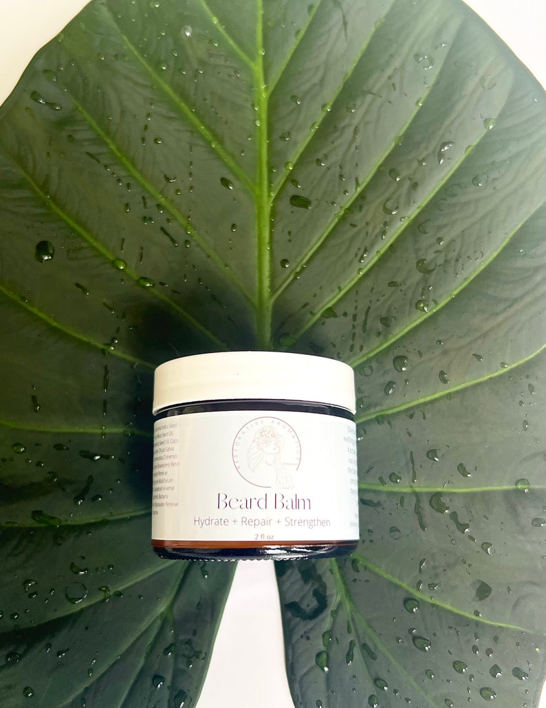 May include: A brown glass jar with a white lid containing beard balm. The jar is labeled "Beard Balm" and "Hydrate + Repair + Strengthen" with a 2 fl oz size. The jar is sitting on a green leaf with water droplets.