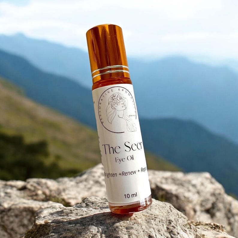 May include: A small, clear glass bottle with a gold cap and a white label. The label says "The Secret Eye Oil" and "Brighten + Renew + Repair". The bottle is sitting on a rock with a mountain range in the background.