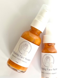 May include: Two amber glass bottles with white pump tops and labels. The labels feature a stylized illustration of a woman with flowers in her hair. The text on the labels reads "Revitalizing Face Serum", "Astaxanthin + CoQ10 + Vitamin C", and "1 fl oz" on the larger bottle and "5 fl oz" on the smaller bottle.