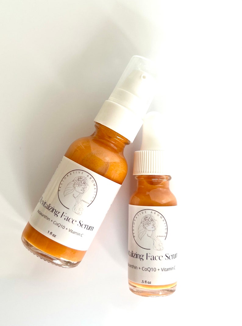 May include: Two amber glass bottles with white pump tops and labels. The labels feature a stylized illustration of a woman with flowers in her hair. The text on the labels reads "Revitalizing Face Serum", "Astaxanthin + CoQ10 + Vitamin C", and "1 fl oz" on the larger bottle and "5 fl oz" on the smaller bottle.