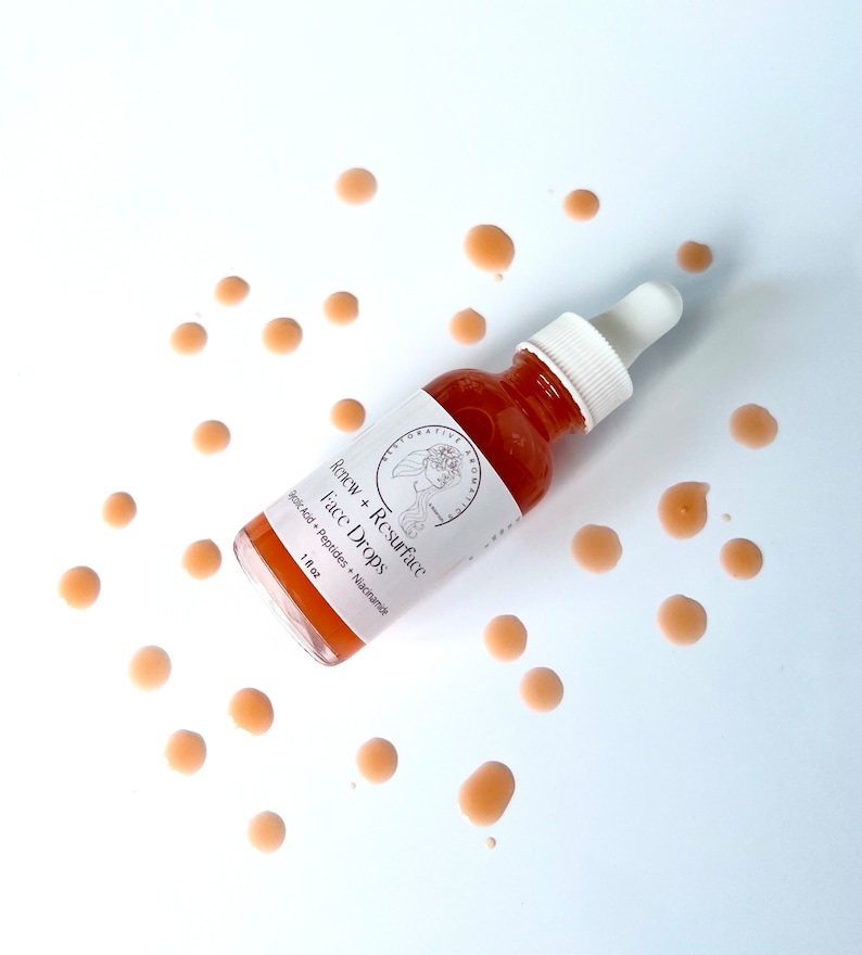 Może przedstawiać: Przezroczysta szklana butelka z kroplomierzem wypełniona jasno pomarańczowym płynem. Butelka jest oznaczona etykietą "Renew + Resurface Face Drops Hyaluronic Acid + Peptides + Niacinamide 1 fl oz". Butelka jest otoczona małymi, jasno pomarańczowymi kroplami na białym tle.