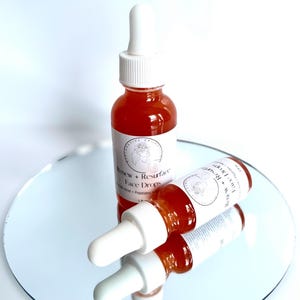 Może przedstawiać: Dwie bursztynowe butelki ze szklanymi pipetkami wypełnione jasnopomarańczowym płynem. Butelki są oznaczone etykietą "Renew + Resurface Face Drops".